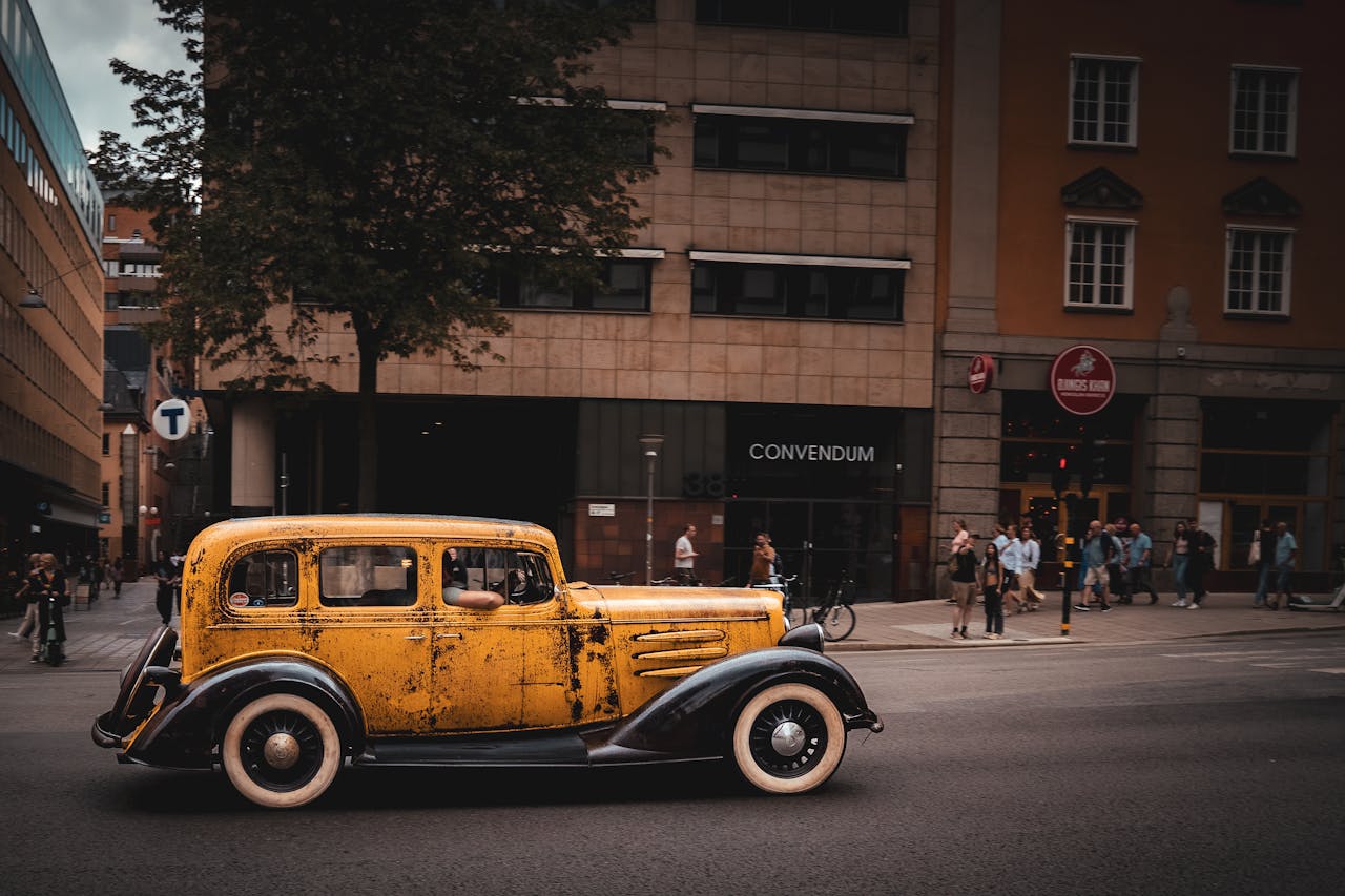 menu-02 Classic yellow vintage car driving on an urban street with city architecture in the background.